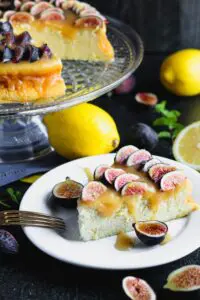 Closeup photo of a slice of lemon yogurt cake topped with honey caramel and quartered figs on a white plate with a crystal cake pedestal with the yogurt cake in the background along with lemons and figs