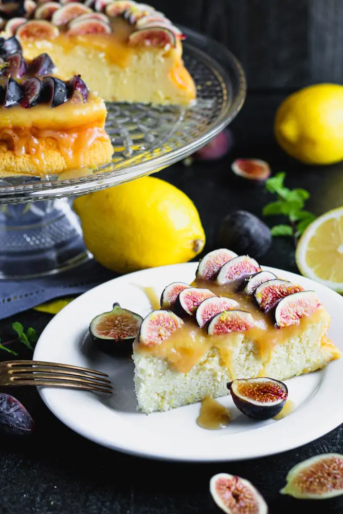 Closeup photo of a slice of lemon yogurt cake topped with honey caramel and quartered figs on a white plate with a crystal cake pedestal with the yogurt cake in the background along with lemons and figs