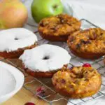 Close up shot of pumpkin donuts with a heavy layer of cream cheese glaze on a cooling rack with more donuts before icing and apples in the background