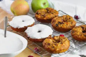 Close up shot of pumpkin donuts with a heavy layer of cream cheese glaze on a cooling rack with more donuts before icing and apples in the background