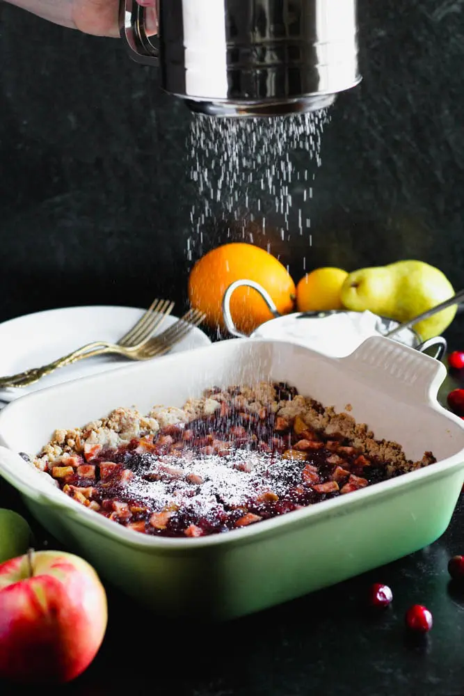 powdered sugar being sifted over the top of uncut pan of cranberry, apple and pear bars in a green baking dish with an apple in the foreground and an orange, pear and lemon in the background