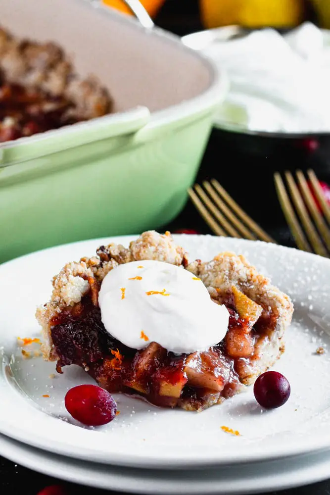 Close up photo of a square piece Cranberry, Apple and Pear Bars with a dollop of whipped cream on a white plate with a green baking dish in the background