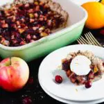a square piece of Cranberry, Apple and Pear bars on a small round white plate with the remaining bars in a green baking dish in the background and pears, apples, and oranges scattered around
