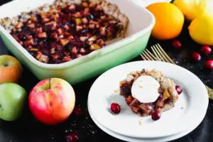 a square piece of Cranberry, Apple and Pear bars on a small round white plate with the remaining bars in a green baking dish in the background and pears, apples, and oranges scattered around