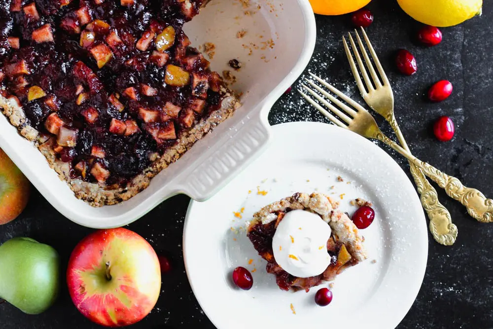 a square piece of Cranberry, Apple and Pear bars on a small round white plate with the remaining bars in a green baking dish in the background and pears, apples, and oranges scattered around