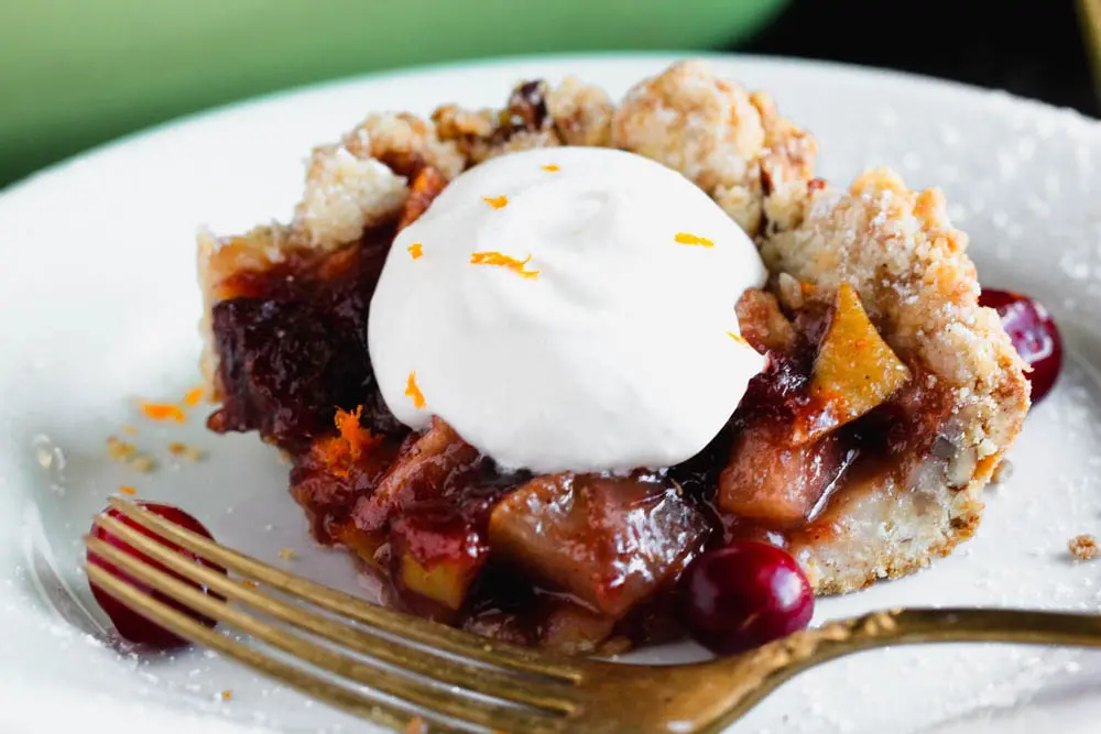 Close up photo of a square piece Cranberry, Apple and Pear Bars with a dollop of whipped cream on a white plate with a green baking dish in the background