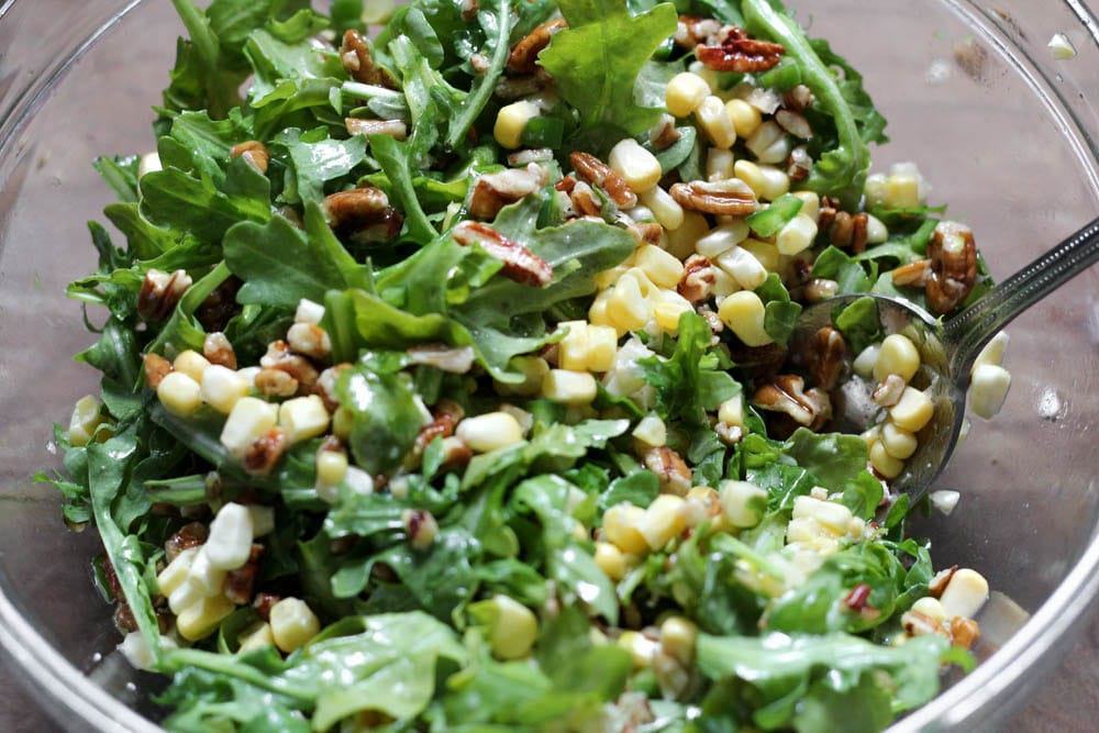 A clear glass bowl filled with a mixture of arugula, brown tree nuts, and yellow corn