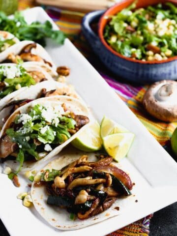 tacos filled with mushrooms and vegetables on a white platter with lime wedges