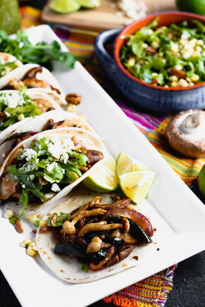 tacos filled with mushrooms and vegetables on a white platter with lime wedges