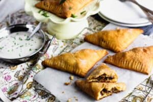 golden brown triangular beef stuffed pastries beside a small metal bowl of white sauce garnished with green herbs
