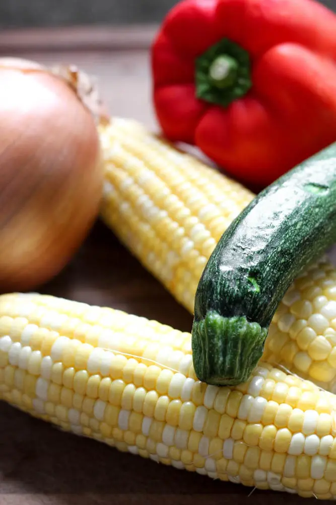 two cobs of corn, a zucchini squash, a red bell pepper, and a yellow onion on a wooden board
