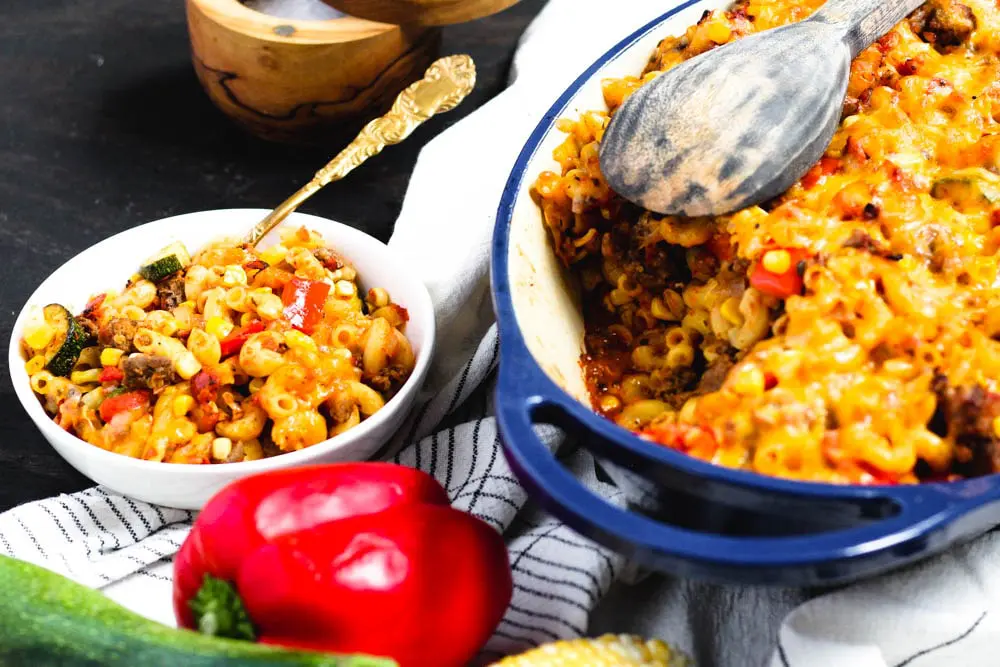 a pasta, squash, corn, and red pepper casserole in a blue baking dish with a serving of the casserole in a bowl to the side