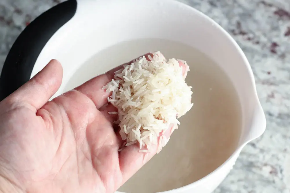 a white bowl filled with rice soaking in water and a hand filled with rice grains from the bowl