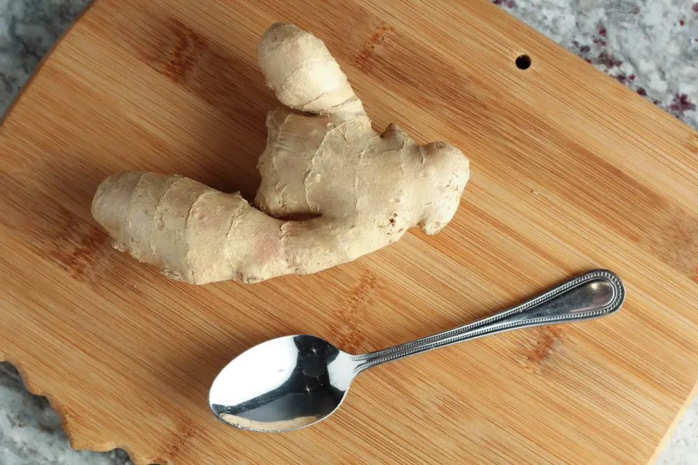 piece of fresh ginger sitting on a bamboo wooden cutting board next to a silver spoon
