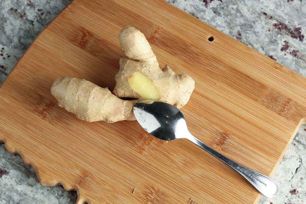piece of ginger on a bamboo cutting board with a silver spoon peeling off skin of ginger
