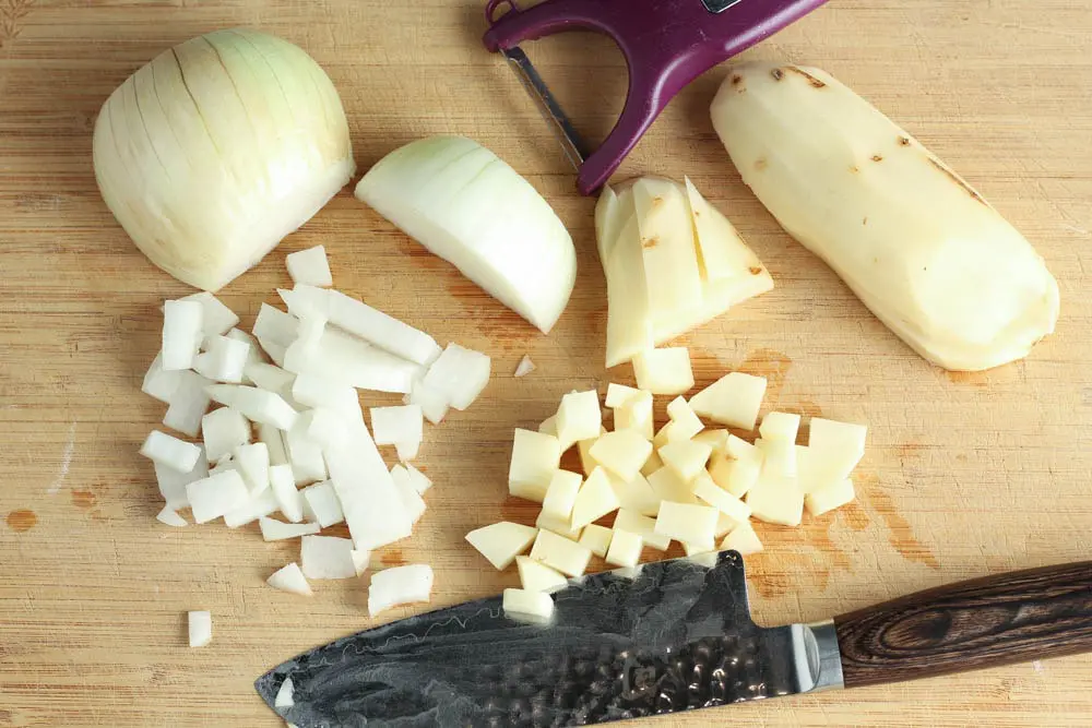 Half a yellow onion with onion diced into quarter inch pieces and a peeled russet potato half with quarter inch diced potatoes with a purple Y vegetable peeler and a silver knife with a dark wooden handle all on a bamboo cutting board