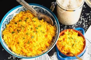 small blue bowl filled with orange rice sitting next to a large decorative blue and white bowl with orange rice and a wooden spoon beside a package of Sazon seasoning