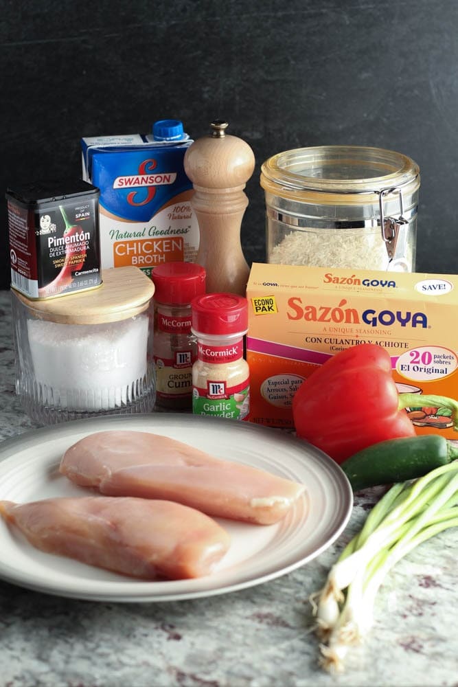 ingredients for easy spanish chicken and rice including smoked paprika, salt, chicken broth, black pepper, garlic powder, cumin, Sazon seasoning, rice, red bell pepper, green jalapeno, green onions, and raw chicken breasts