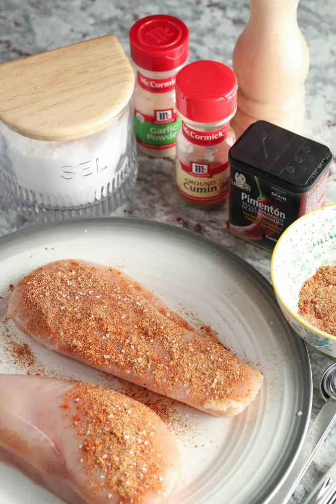 white round plate with a grey rim with raw chicken breasts seasoned with spices in the background including garlic powder, salt, powder, paprika and cumin