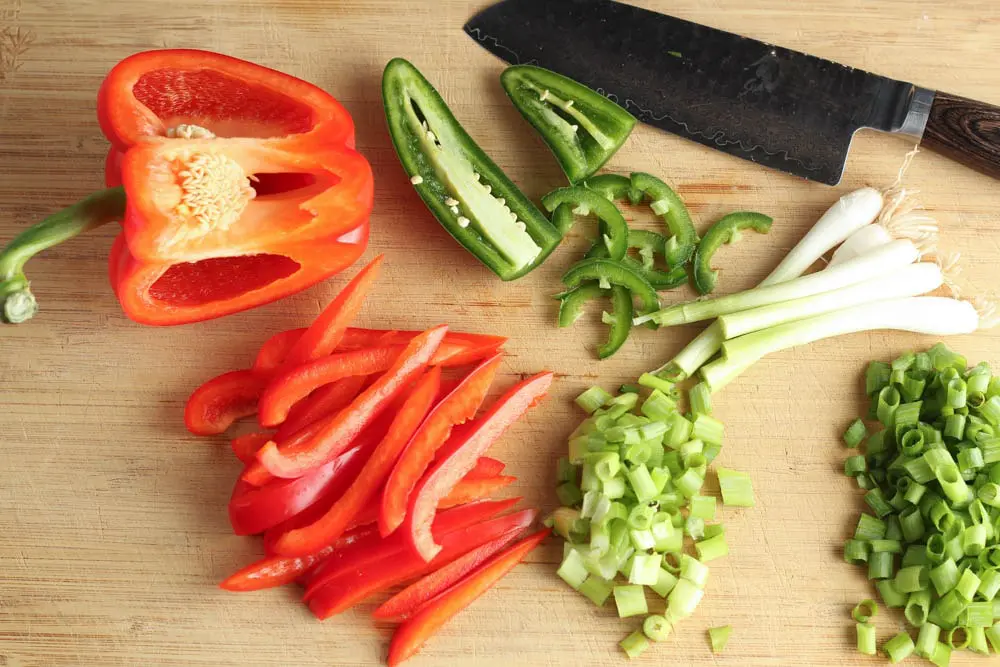 wooden cutting board with a silver knife and chopped green onions, sliced jalapenos and red bell pepper strips