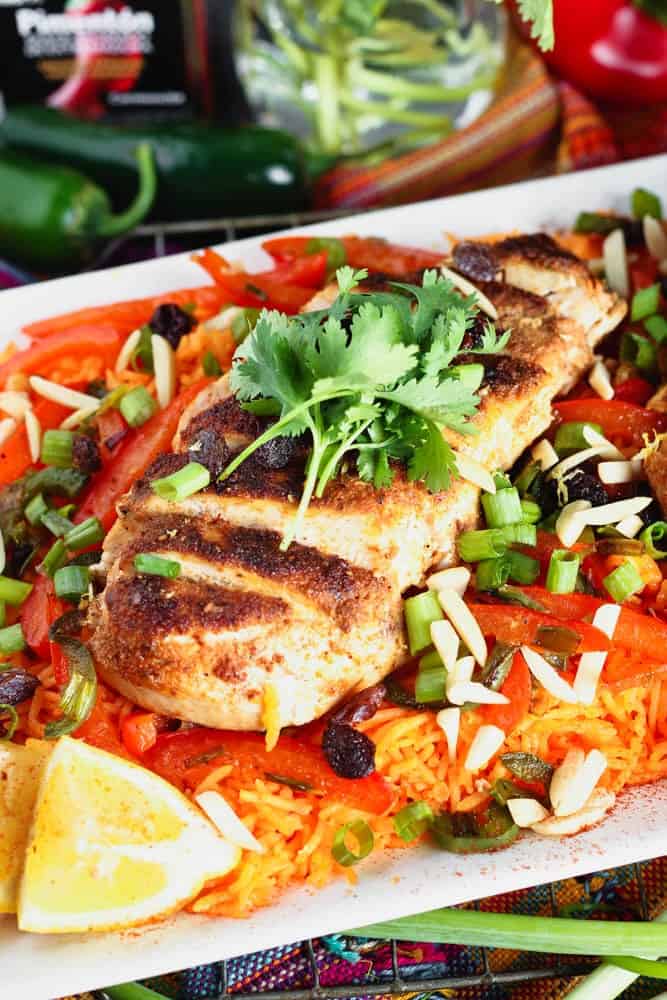 white rectangular plate filled with orange rice topped with strips of red bell pepper, green onion pieces, sliced chicken breast and cilantro with a lemon wedge