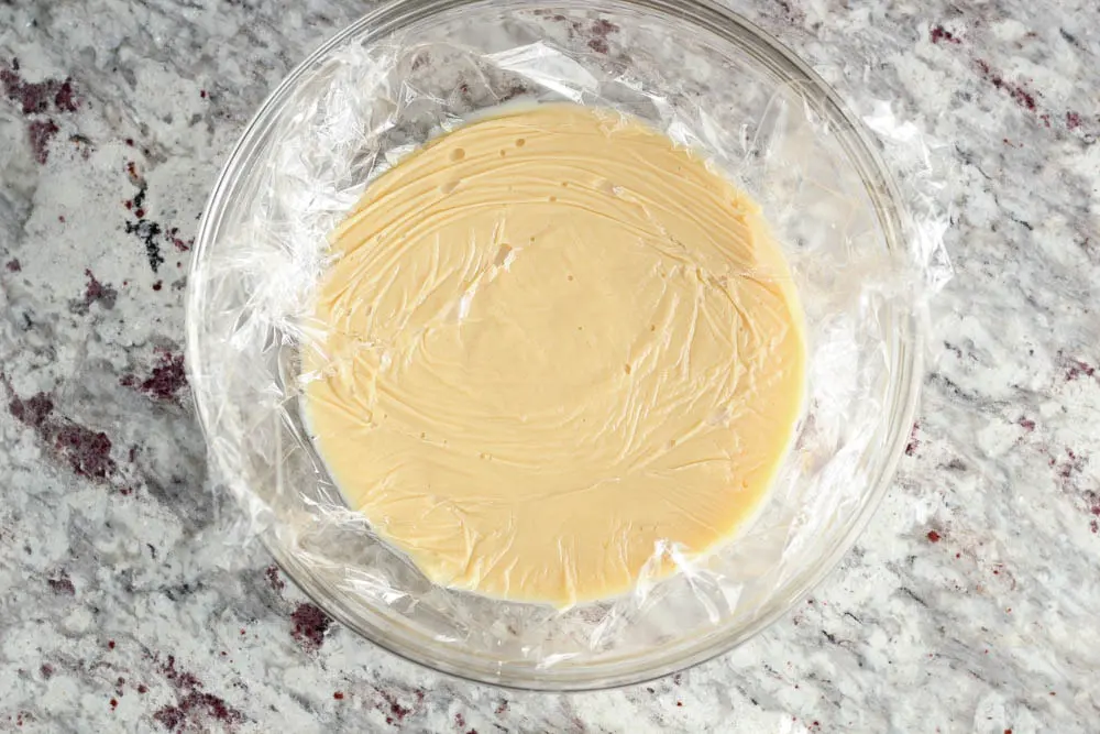 overhead view of pale yellow vanilla pudding in a clear glass mixing bowl with plastic wrap pressed all the way to the surface of the pudding