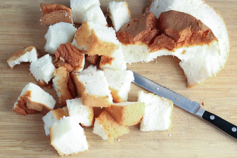 brown and white angel food cake on a wooden cutting board being cut into one inch pieces by a silver serrated knife