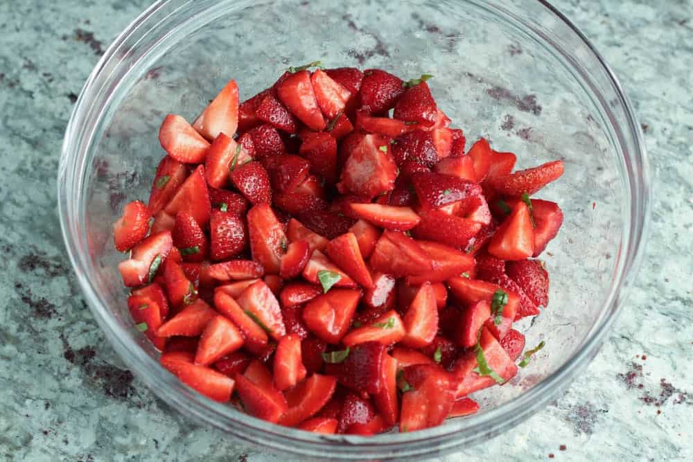 red strawberry pieces in a clear glass mixing bowl with small bits of green basil