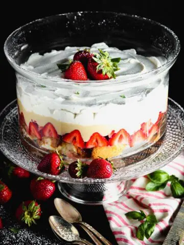 Clear round glass dish filled with layers of angel food cake, bright red strawberries, pale yellow vanilla pudding and white whipped cream topped with three whole strawberries on a clear glass pedestal with a pink and white napkin and four antique silver spoons