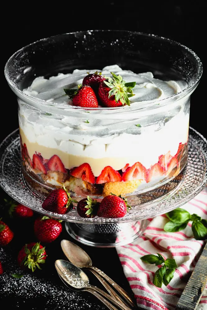 Clear round glass dish filled with layers of angel food cake, bright red strawberries, pale yellow vanilla pudding and white whipped cream topped with three whole strawberries on a clear glass pedestal with a pink and white napkin and a brown wooden spoon