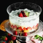 Clear round glass dish filled with layers of angel food cake, bright red strawberries, pale yellow vanilla pudding and white whipped cream topped with three whole strawberries on a clear glass pedestal with a pink and white napkin and a brown wooden spoon