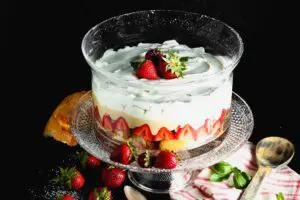 Clear round glass dish filled with layers of angel food cake, bright red strawberries, pale yellow vanilla pudding and white whipped cream topped with three whole strawberries on a clear glass pedestal with a pink and white napkin and a brown wooden spoon