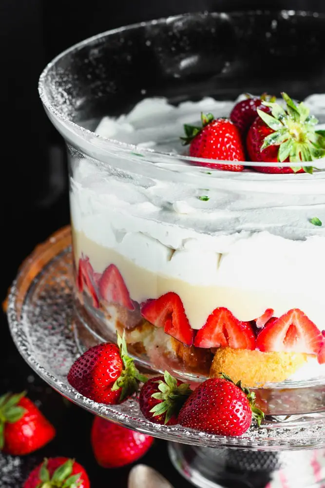 Clear round glass dish filled with layers of angel food cake, bright red strawberries, pale yellow vanilla pudding and white whipped cream topped with three whole strawberries on a clear glass pedestal