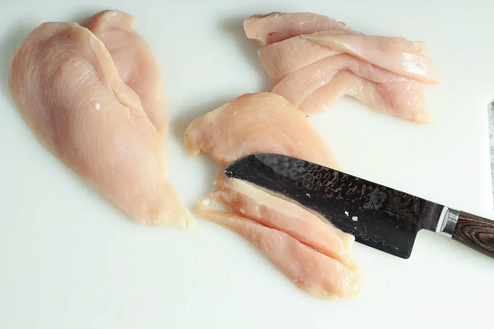 raw chicken breast on a white cutting board being sliced into thin strips