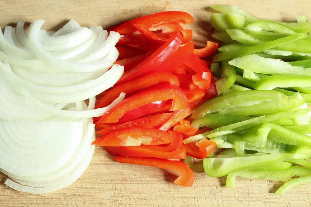 white onion and bright red and green bell peppers thinly sliced on a light wooden cutting board