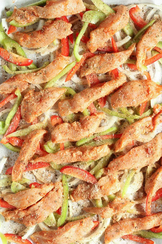 raw chicken and uncooked onions and peppers spread across a baking sheet lined with parchment paper sprinkled with herbs and spices