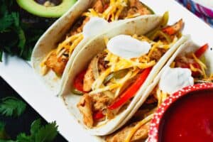 Three prepared chicken fajitas tacos with slices of chicken, peppers, onions, shredded cheese and a dollop of sour cream with a bowl of red salsa and fresh green avocadoes on the side
