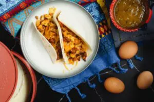 Two chorizo and potato breakfast tacos filled with orange chorizo, yellow scrambled eggs and chunks of potato and onion served in a flour tortilla on a white plate and a festive Mexican blanket backdrop