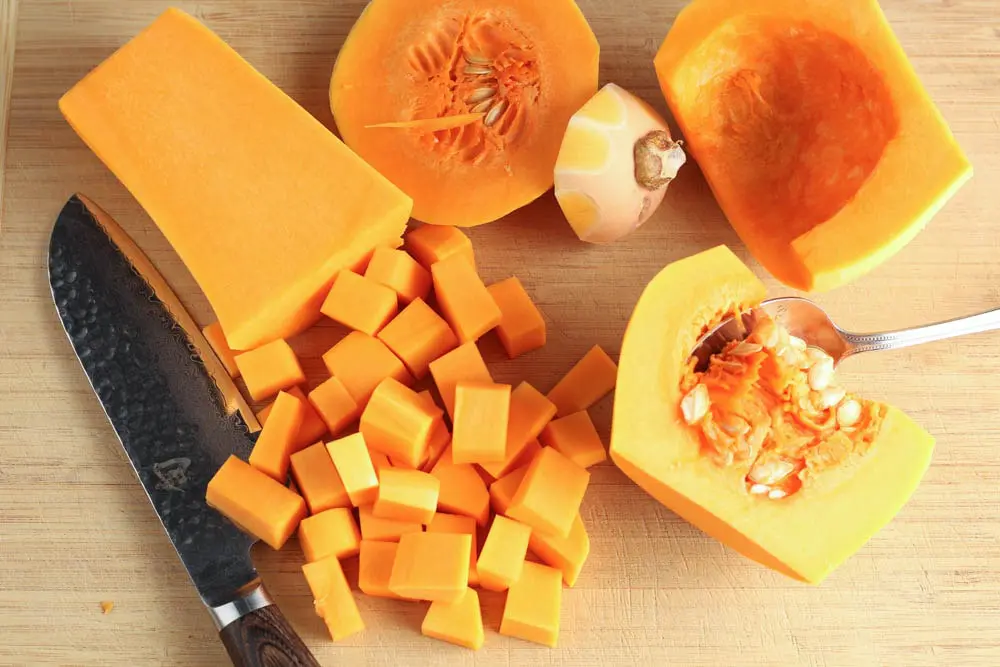Light wooden cutting board with a split bright orange butternut squash, diced butternut squash, a spoon for scooping out the squash pulp and a chef's knife