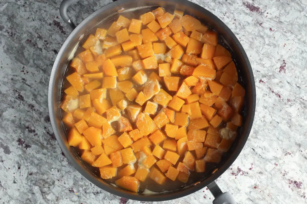 a large black high rimmed saute pan filled with bright orange butternut squash cut into one inch cubes and covered with chicken stock