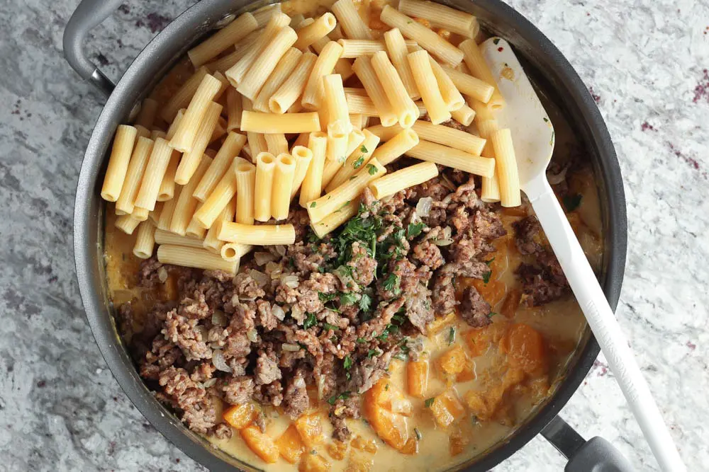 Large high rimmed saute pan filled with complete recipe before mixing together; one section contains cooked pasta, one the cooked sausage and onions mixture and last the bright orange cooked butternut squash