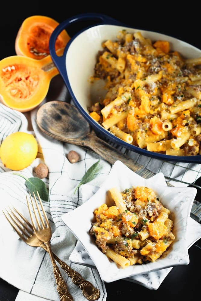 White star shaped bowl filled filled with orange butternut squash cubes, sausage pieces, rigatoni and fleck of white parmesan cheese and green chopped parsley and a blue casserole dish in the background filled with the recipe