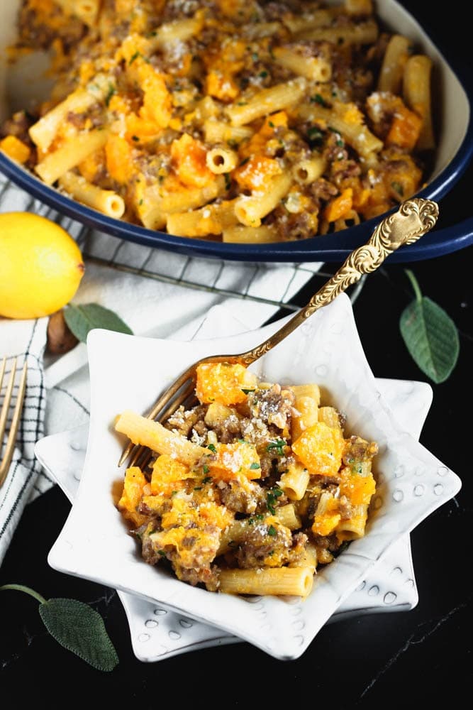 White star shaped bowl filled filled with orange butternut squash cubes, sausage pieces, rigatoni and fleck of white parmesan cheese and green chopped parsley and a blue casserole dish in the background filled with the recipe