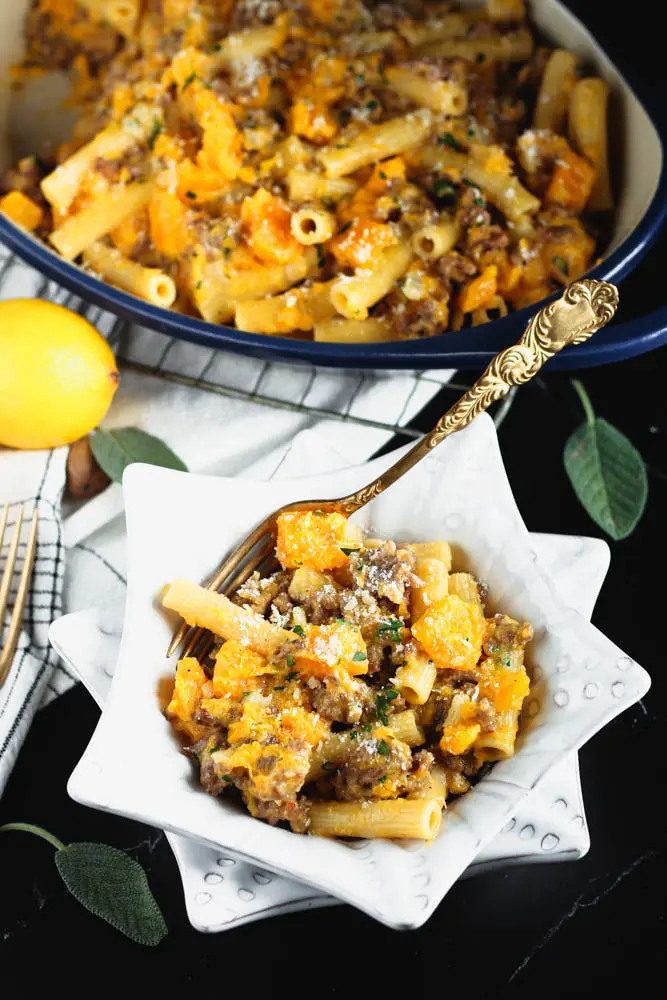 White star shaped bowl filled filled with orange butternut squash cubes, sausage pieces, rigatoni and fleck of white parmesan cheese and green chopped parsley and a blue casserole dish in the background filled with the recipe