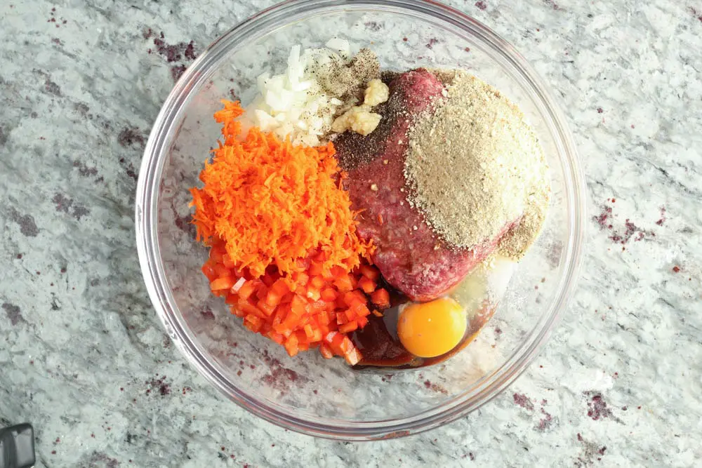 large glass bowl filled with meatloaf ingredients prior to mixing including grated orange carrot, diced red bell pepper and white onion, ground beef, breadcrumbs and a raw egg