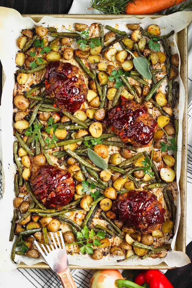 Small round meatloaf portions topped with rich red barbecue sauce sitting on a golden baking sheet topped with parchment paper and filled with golden yellow potatoes and green parsley and sage leaves