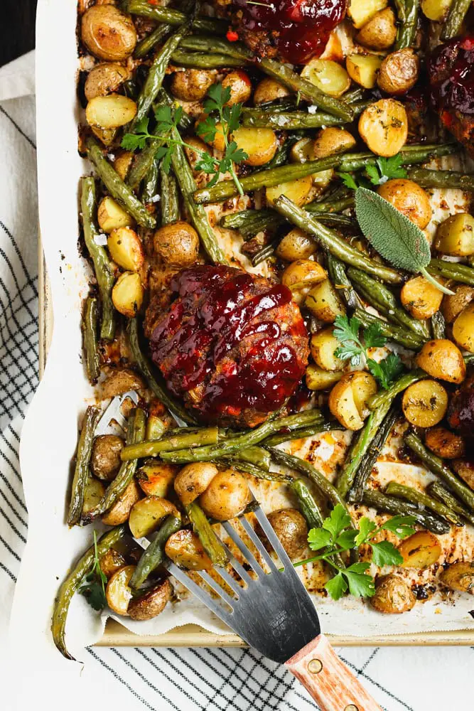 Small round meatloaf portions topped with rich red barbecue sauce sitting on a golden baking sheet topped with parchment paper and filled with golden yellow potatoes and green parsley and sage leaves