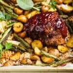 Small round meatloaf portions topped with rich red barbecue sauce sitting on a golden baking sheet topped with parchment paper and filled with golden yellow potatoes and green parsley and sage leaves