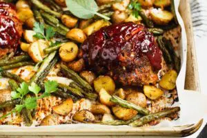 Small round meatloaf portions topped with rich red barbecue sauce sitting on a golden baking sheet topped with parchment paper and filled with golden yellow potatoes and green parsley and sage leaves