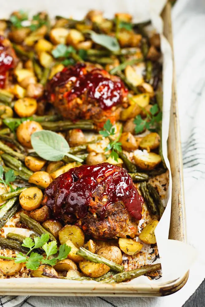 Small round meatloaf portions topped with rich red barbecue sauce sitting on a golden baking sheet topped with parchment paper and filled with golden yellow potatoes and green parsley and sage leaves